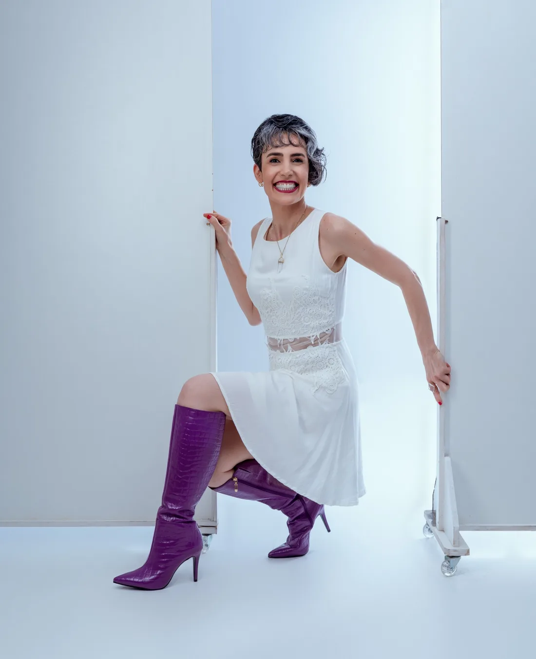 Anaritta Silveira, terapeuta integrativa, agachada entre painéis de estúdio, de vestido branco e botas roxas, sorrindo